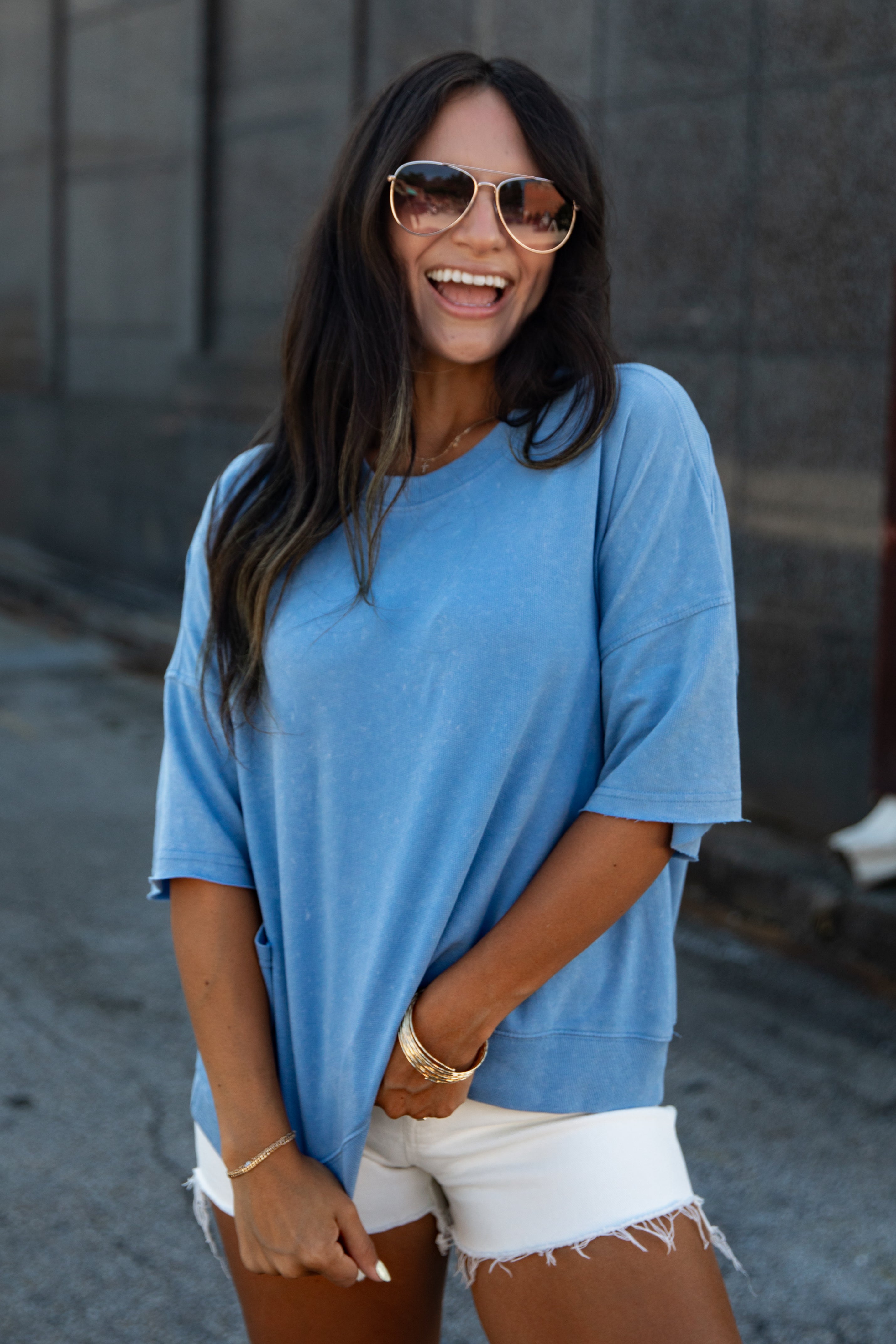A woman wearing a light blue oversized short-sleeve t-shirt and white frayed denim shorts, accessorized with gold-framed sunglasses and gold jewelry, standing outdoors against a gray concrete wall.