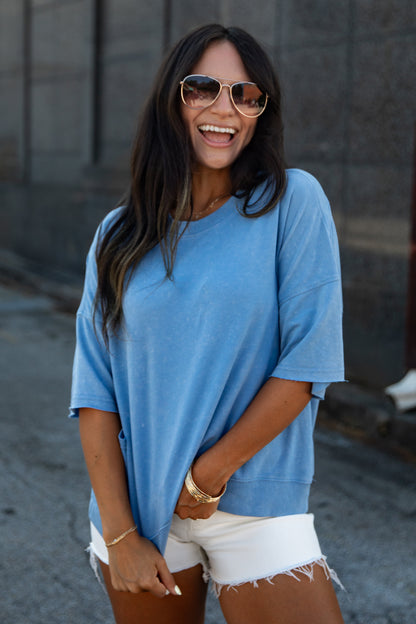 A woman wearing a light blue oversized short-sleeve t-shirt and white frayed denim shorts, accessorized with gold-framed sunglasses and gold jewelry, standing outdoors against a gray concrete wall.