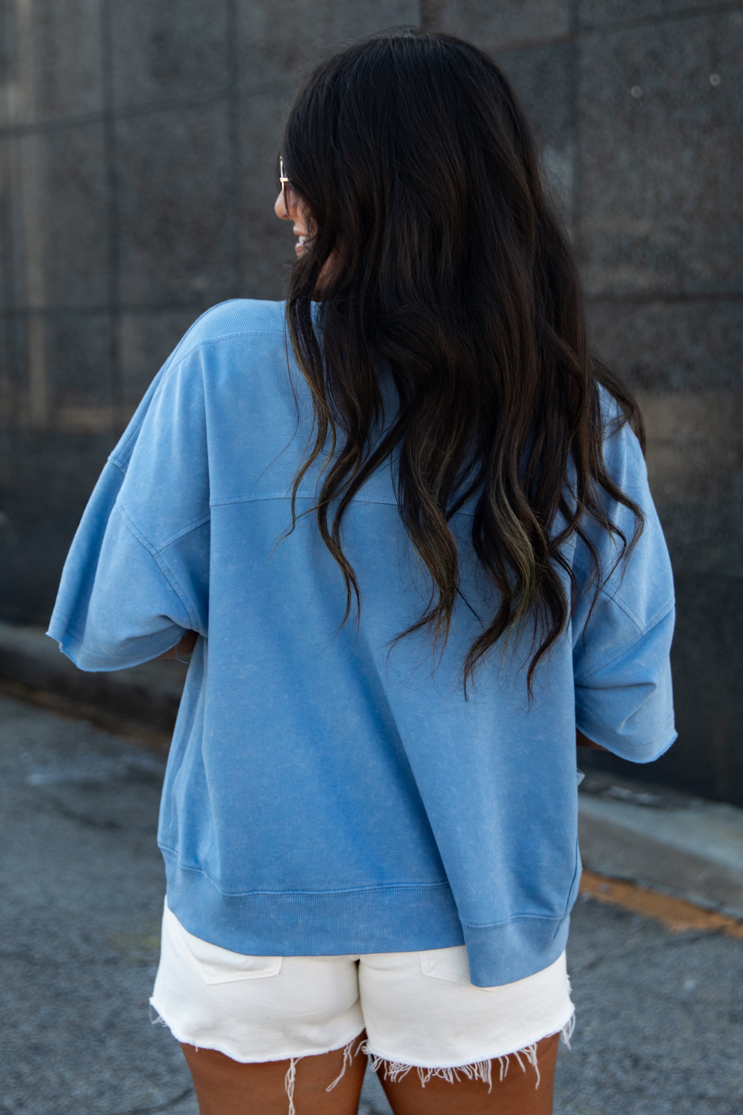 A woman viewed from behind wearing an oversized light blue sweatshirt and white frayed denim shorts, standing against a dark gray stone wall.