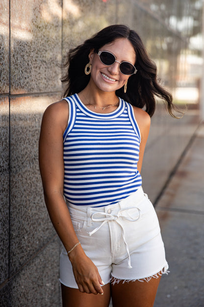 A woman wearing a blue and white striped tank top and white denim shorts stands outdoors against a stone wall.