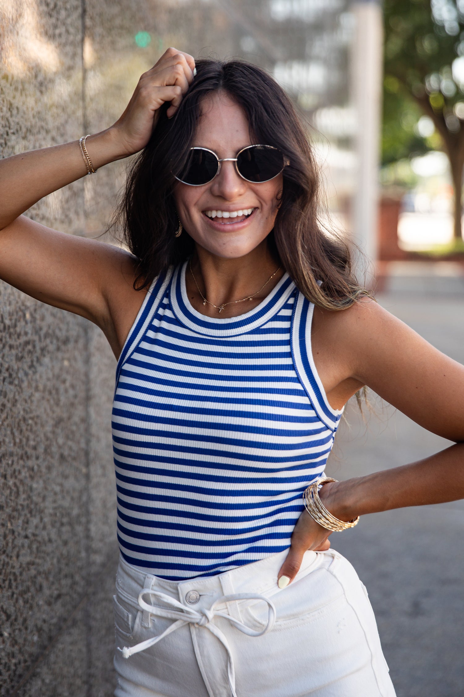 A woman wearing a blue and white striped tank top and white drawstring pants, posing outdoors against a textured wall.