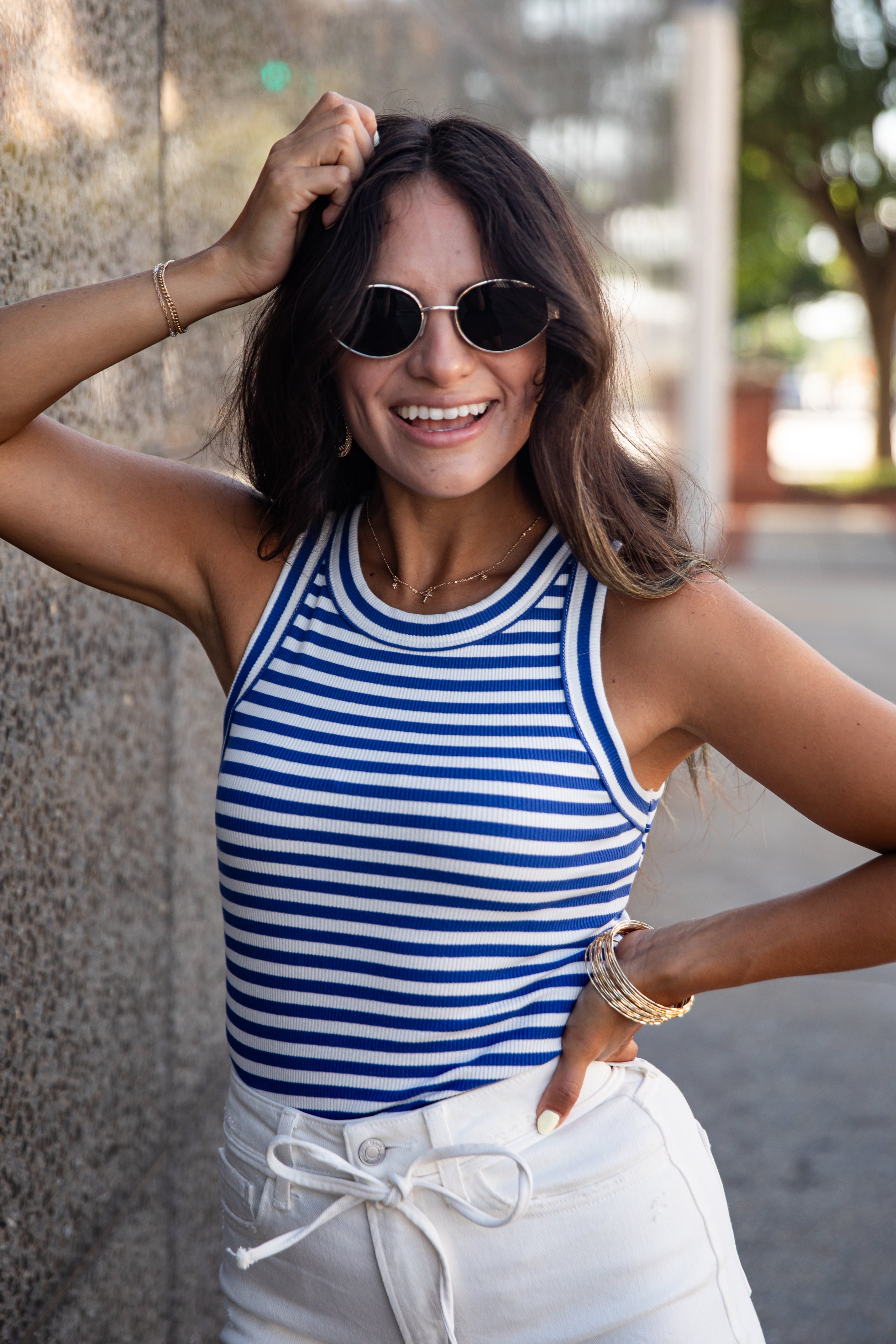 A woman wearing a blue and white striped tank top and white drawstring pants, posing outdoors against a textured wall.