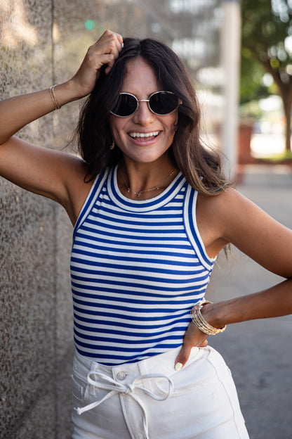 A woman wearing a blue and white striped tank top and white drawstring pants, posing outdoors against a textured wall.