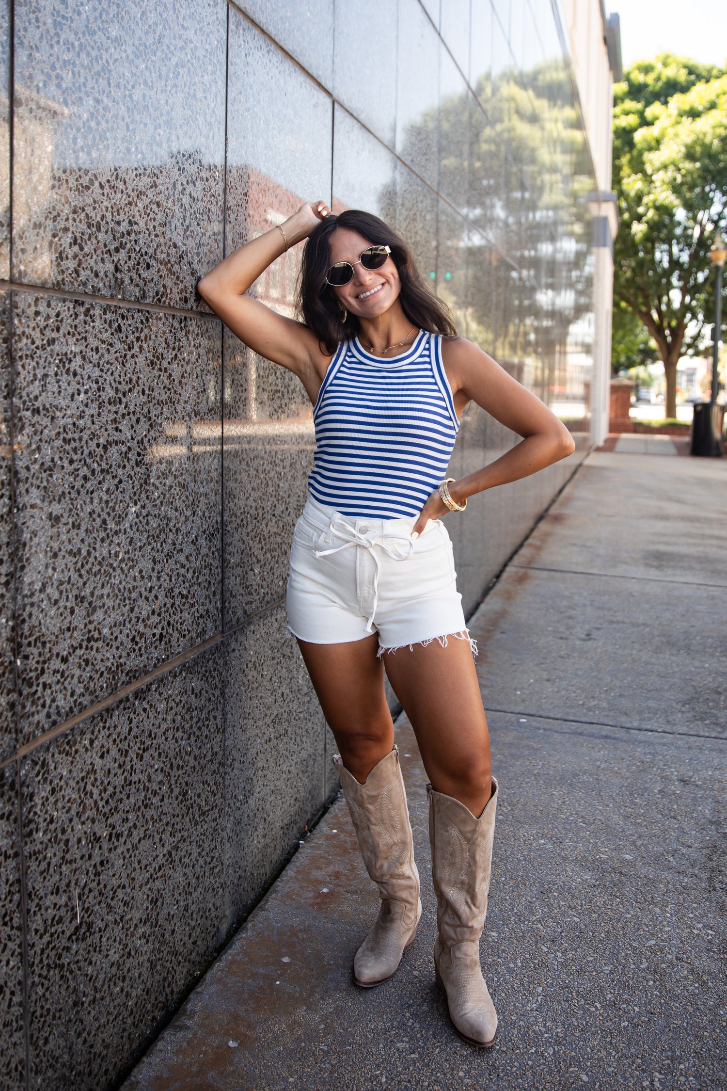 A woman posing on a city sidewalk against a gray stone wall, wearing a blue and white striped tank top, white denim shorts, and beige knee-high boots.