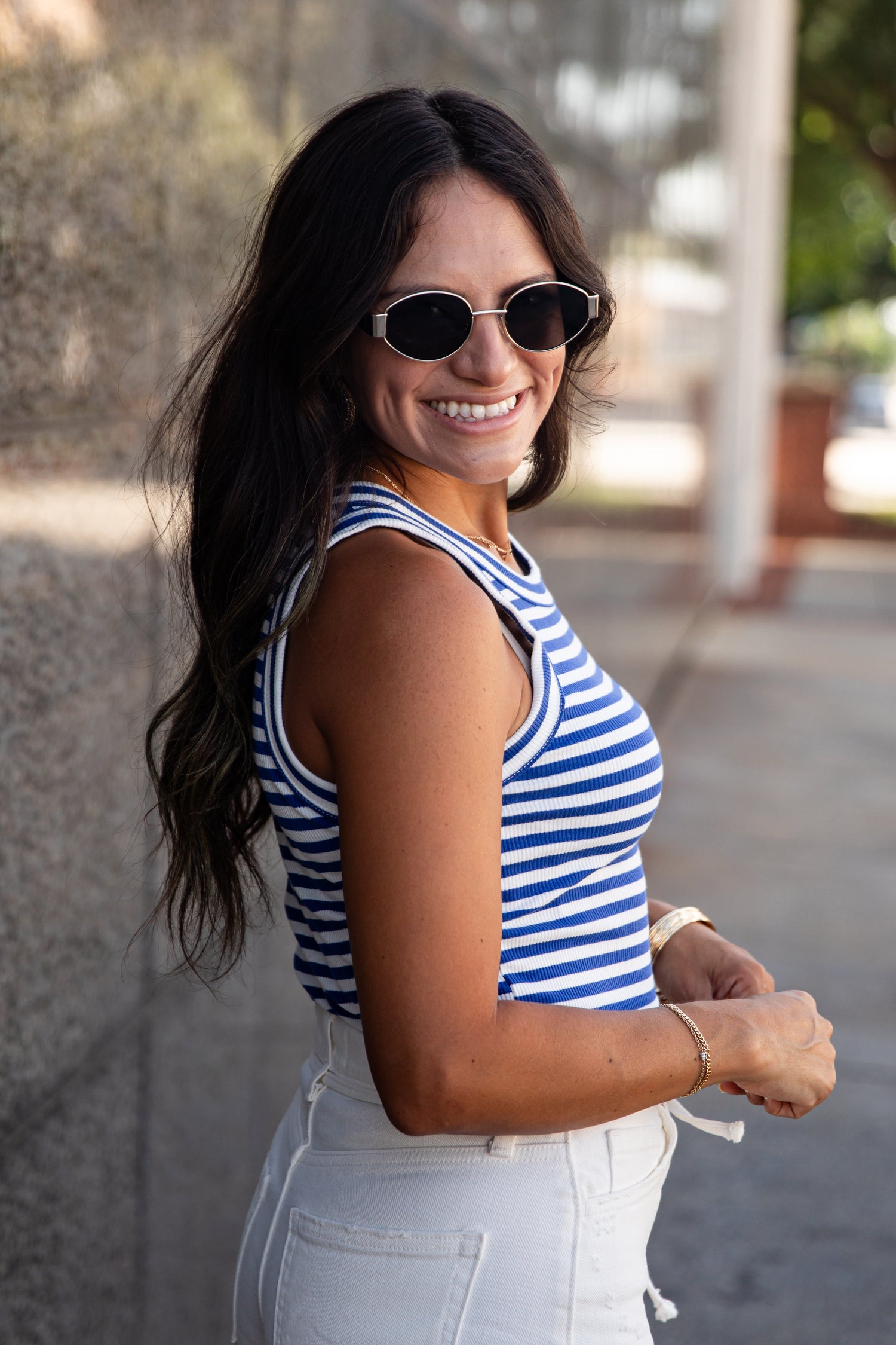 A woman wearing a blue and white striped tank top and white jeans, smiling and looking back over her shoulder in an outdoor setting.