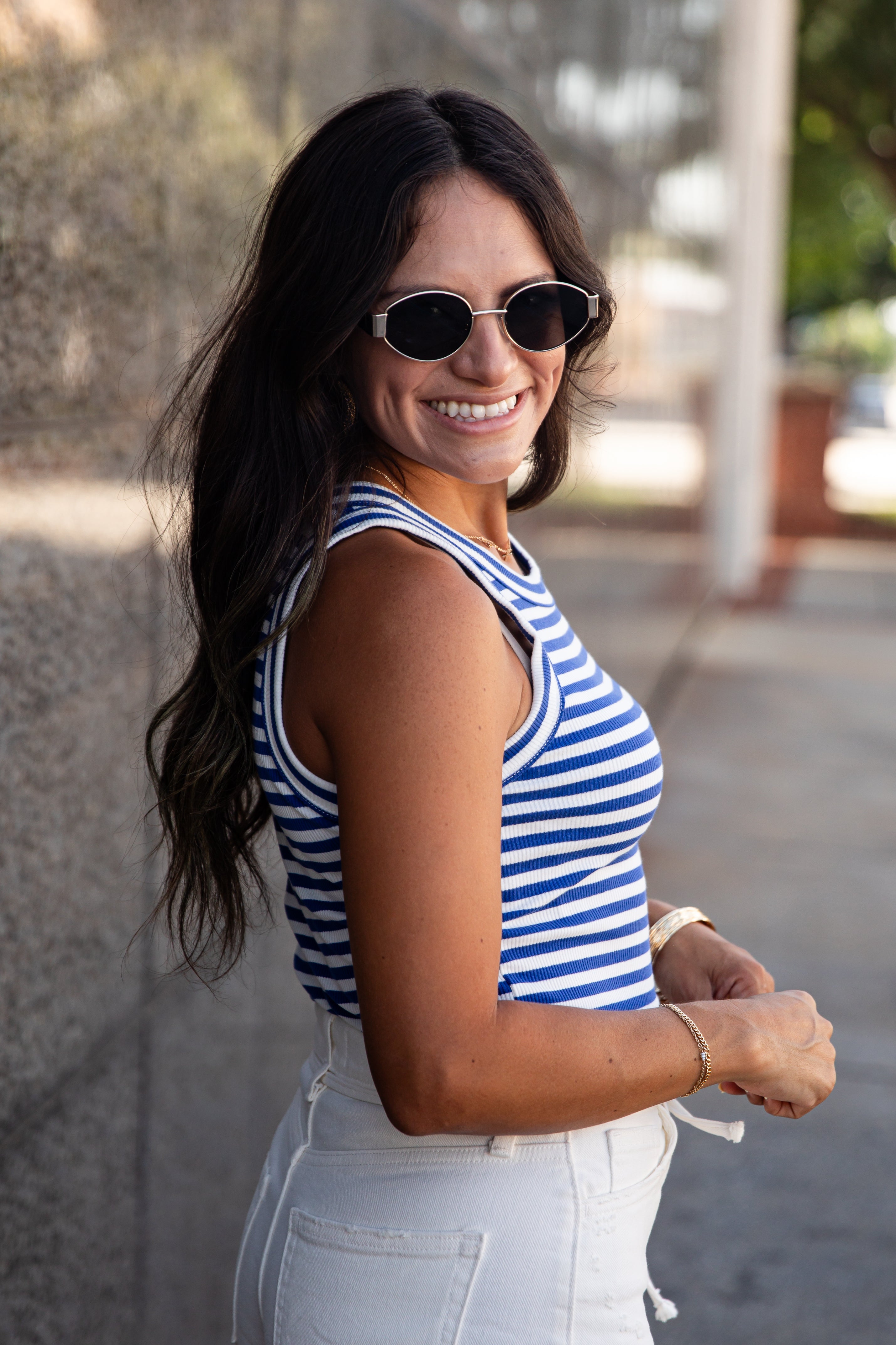 A woman wearing a blue and white striped tank top and white jeans, smiling and looking back over her shoulder in an outdoor setting.