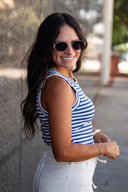 A woman wearing a blue and white striped tank top and white jeans, smiling and looking back over her shoulder in an outdoor setting.