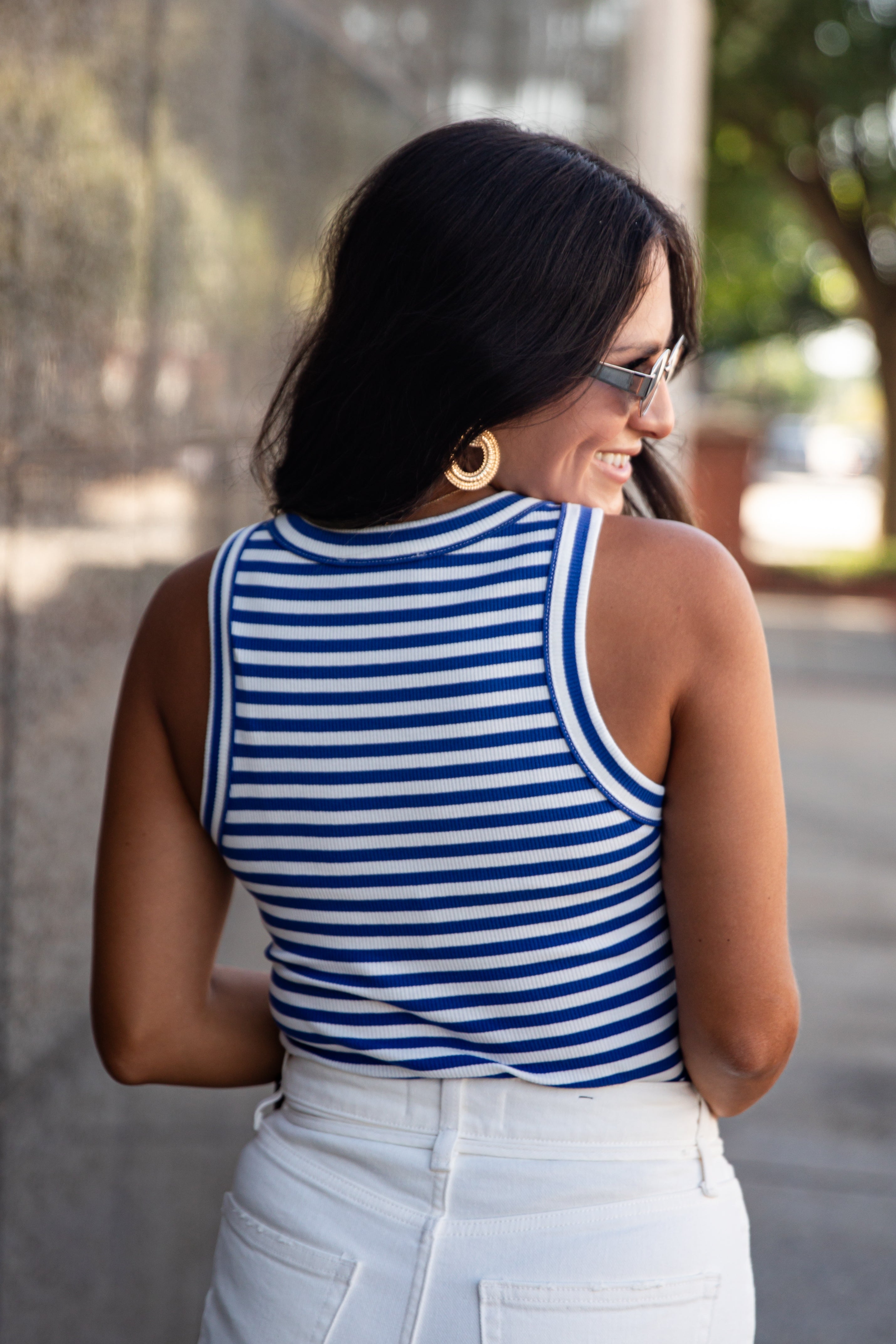 A woman wearing a blue and white striped sleeveless top and white pants, photographed from behind in an outdoor setting.