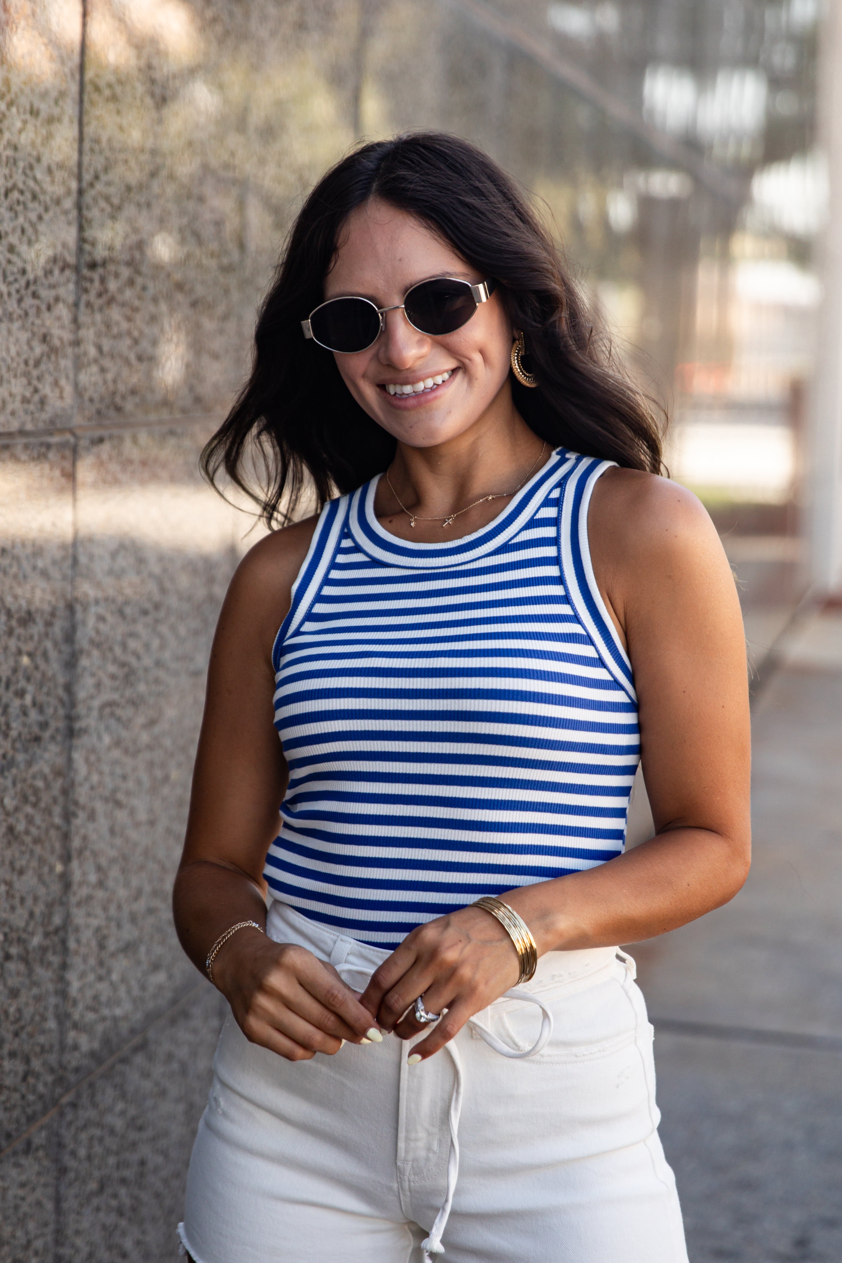 A woman wearing a blue and white horizontally striped tank top and white drawstring shorts, standing outdoors against a stone wall.