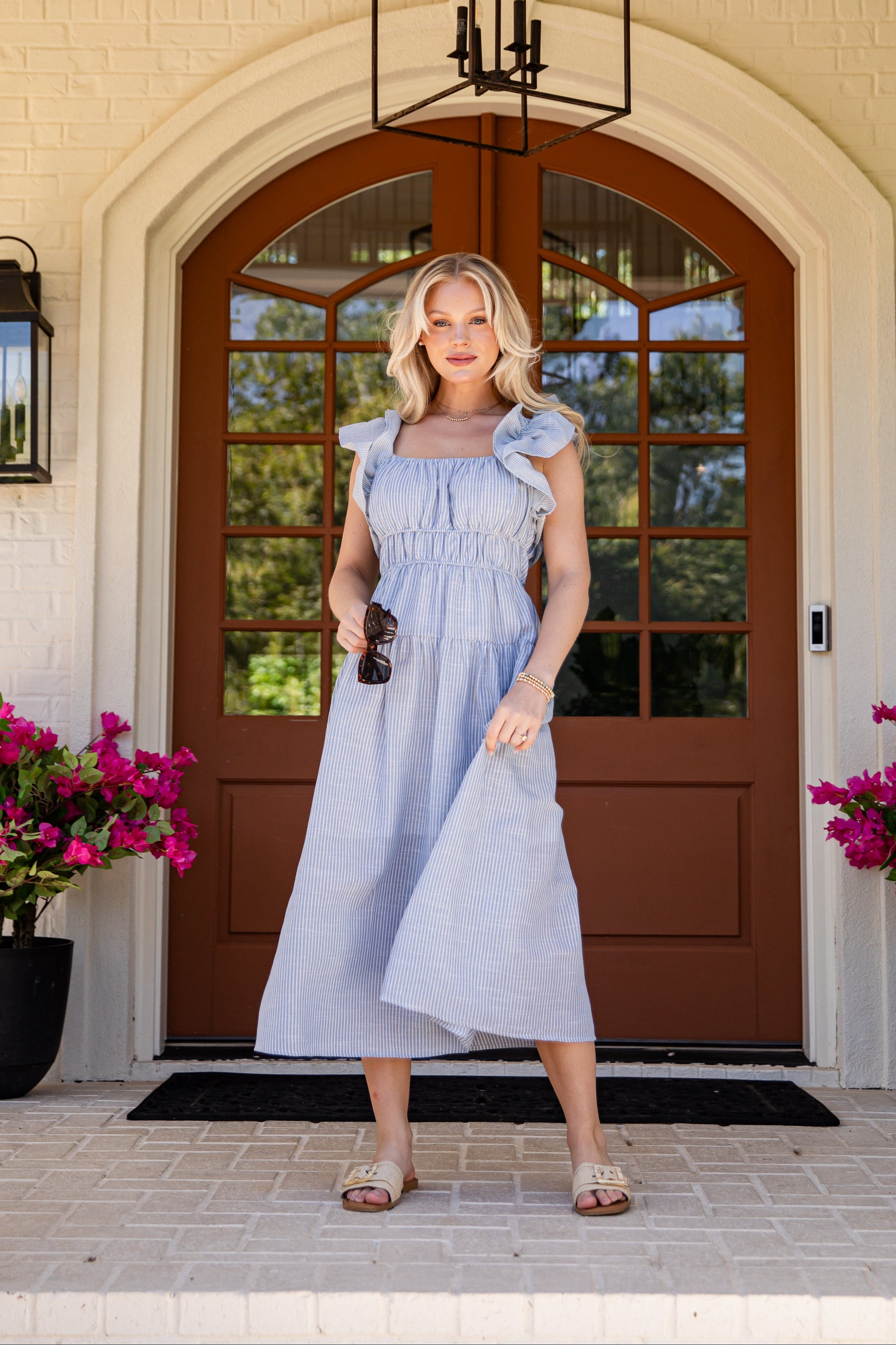 Woman in a light blue dress standing in front of a house entrance with pink flowers on either side.