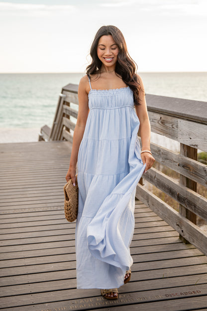 Woman in a light blue dress standing on a wooden pier by the water.