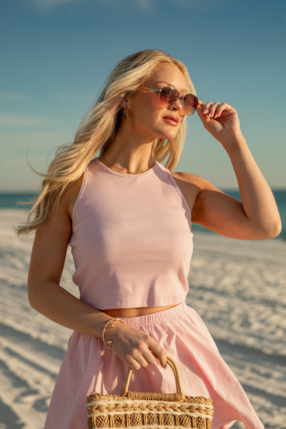 Woman in a pink outfit on a beach with sunglasses and a straw bag.