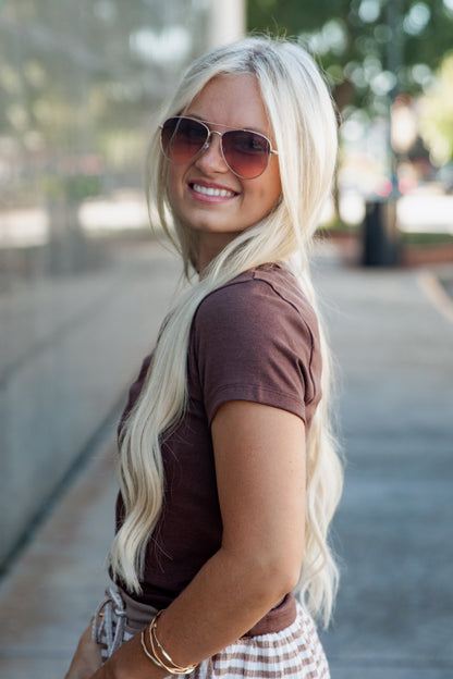 A smiling blonde woman wearing aviator sunglasses, a dark brown short-sleeve t-shirt, and a plaid skirt, photographed outdoors on a blurred sidewalk background.