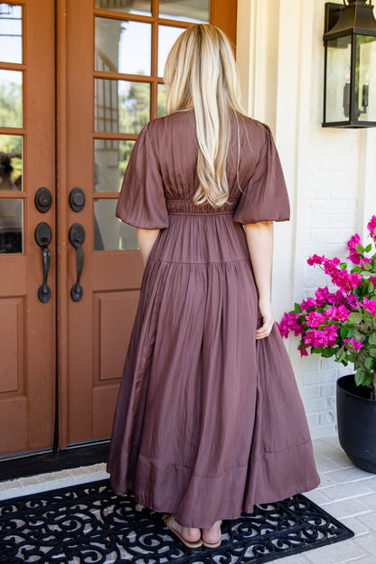 A woman stands with her back to the camera wearing a dusty mauve-brown tiered maxi dress with short puff sleeves, standing in front of a brown wooden front door with a black doormat and pink flowers on the porch.