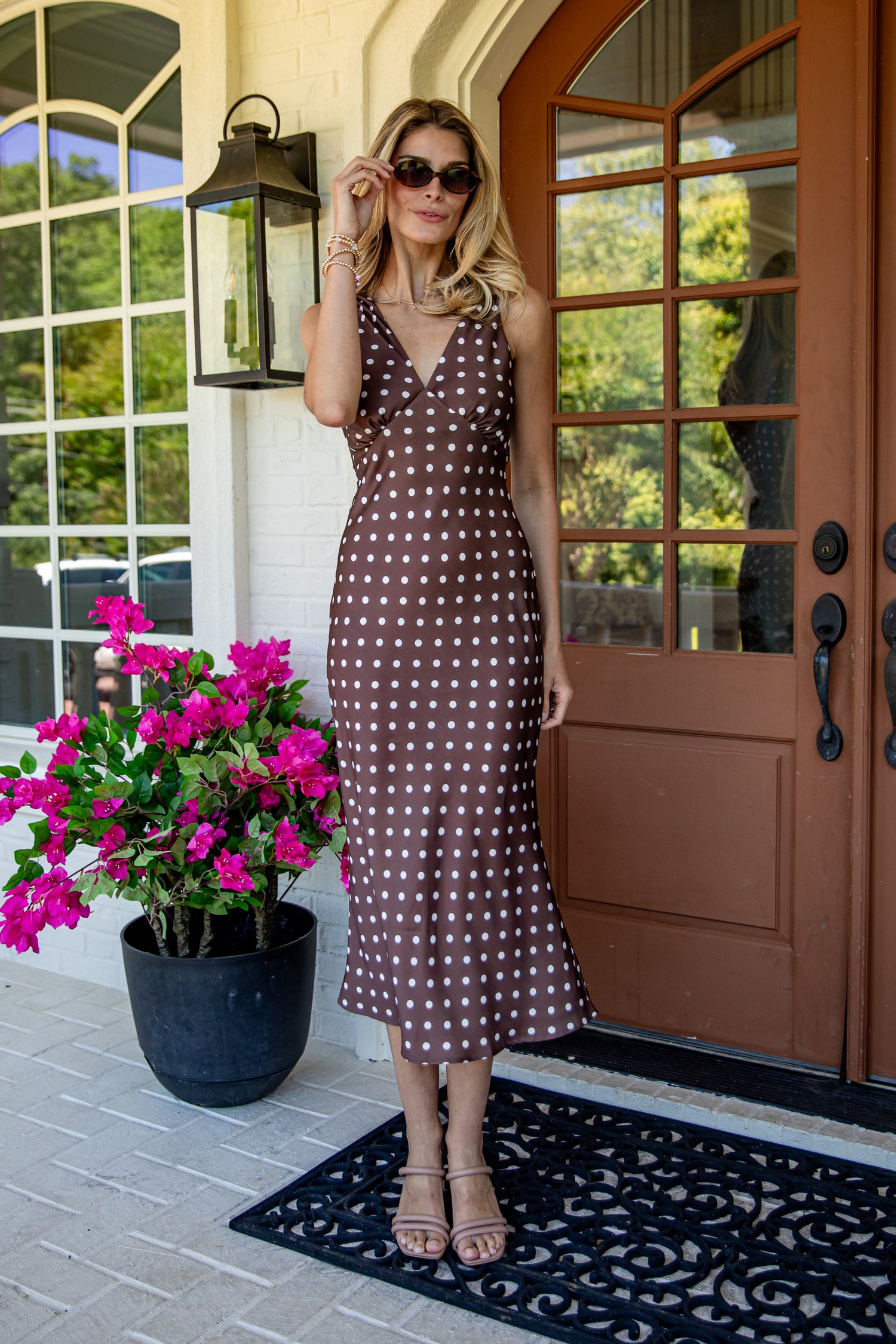A woman wearing a brown and white polka dot midi dress stands on a front porch next to a potted plant with bright pink flowers.