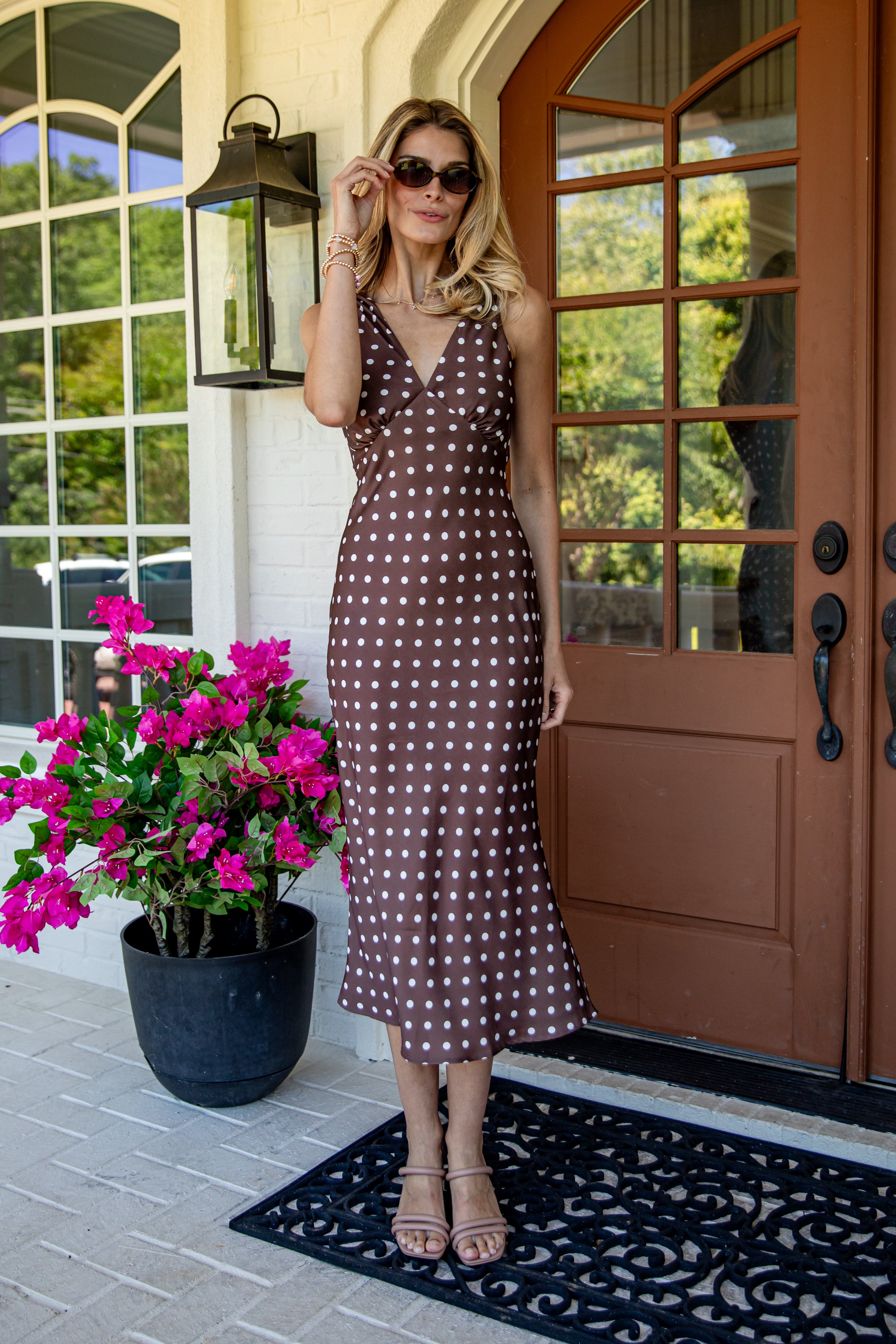 A woman wearing a brown and white polka dot midi dress stands on a front porch next to a potted plant with bright pink flowers.