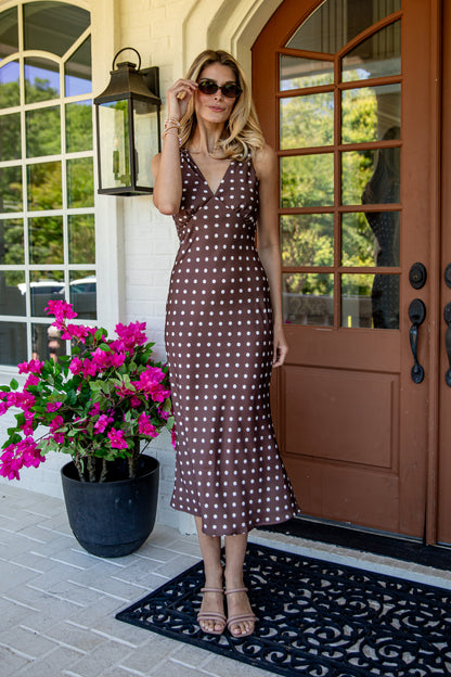 A woman wearing a brown and white polka dot midi dress stands on a front porch next to a potted plant with bright pink flowers.