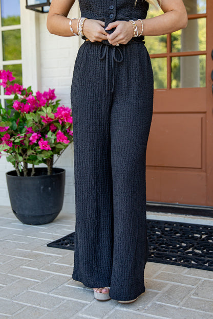 A person wearing dark charcoal gray wide-leg textured pants with a drawstring waist, standing on a tiled porch with a potted pink flower and a brown door in the background.