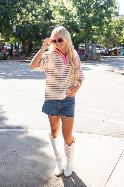 A woman stands on a sunny street wearing a cream and brown horizontally striped short-sleeve top with a pink collar, blue denim shorts, and white cowboy boots.