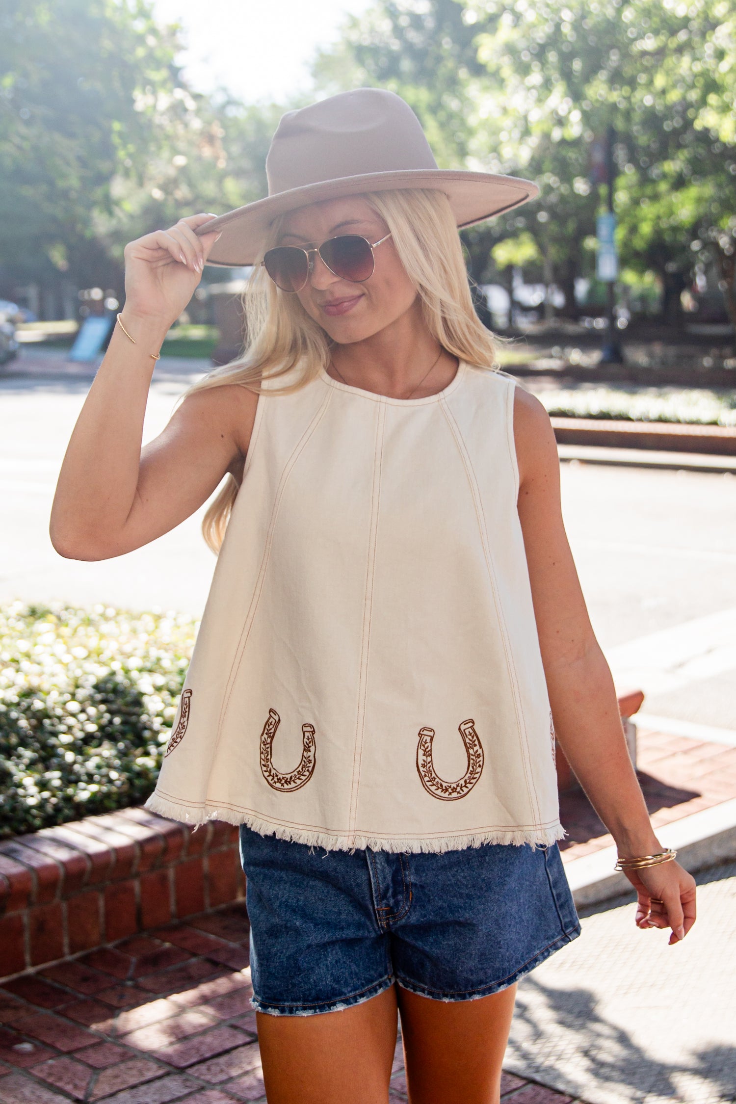 A woman wearing a sleeveless cream-colored top with embroidered horseshoes, blue denim shorts, a beige wide-brim hat, and sunglasses, standing outdoors on a brick sidewalk.