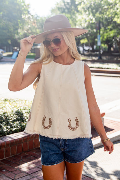 A woman wearing a sleeveless cream-colored top with embroidered horseshoes, blue denim shorts, a beige wide-brim hat, and sunglasses, standing outdoors on a brick sidewalk.