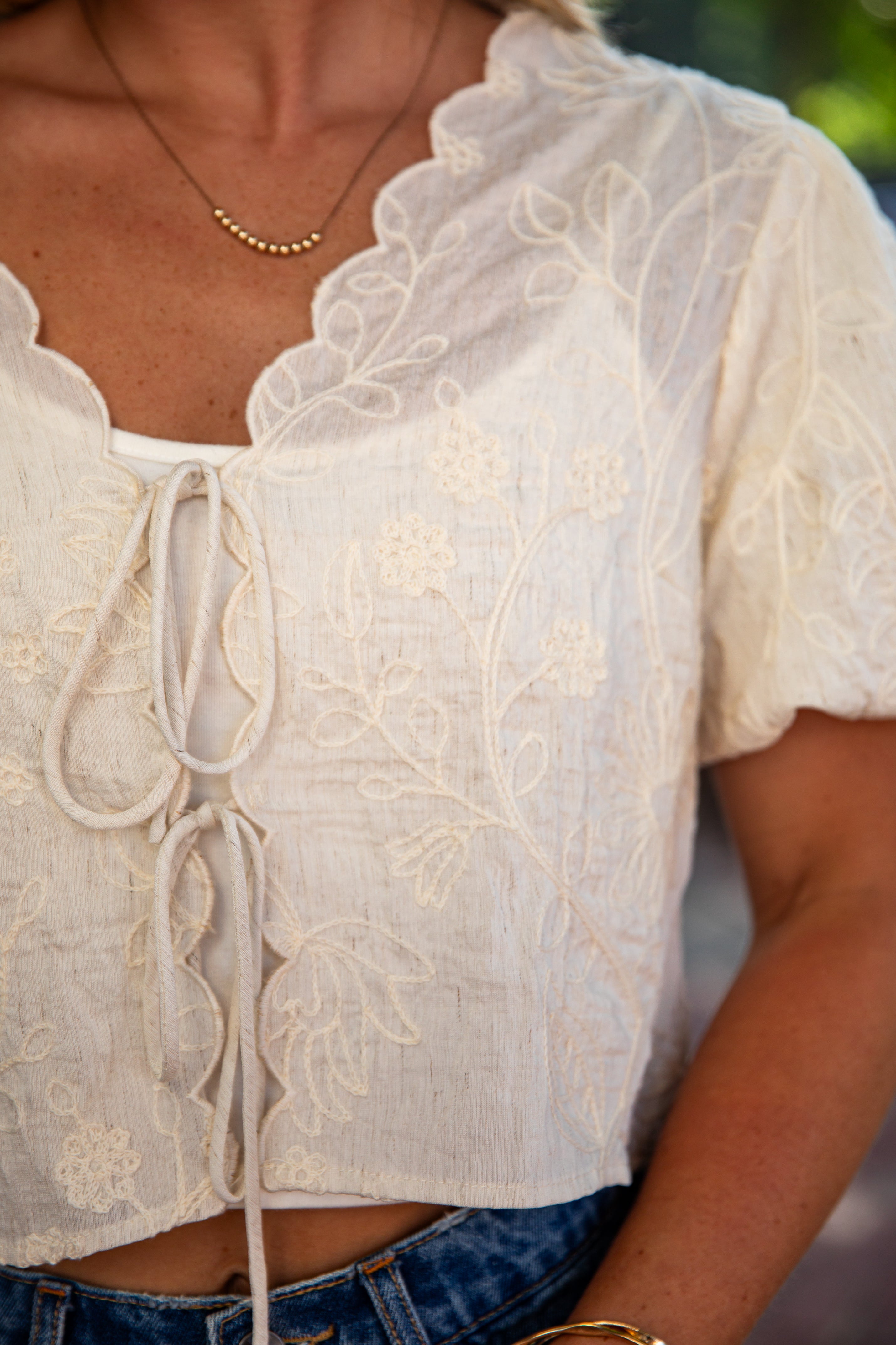 A close-up of a person wearing a cream-colored short-sleeve lace top with a tie-front closure and floral embroidery, paired with blue jeans.