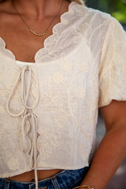A close-up of a person wearing a cream-colored short-sleeve lace top with a tie-front closure and floral embroidery, paired with blue jeans.