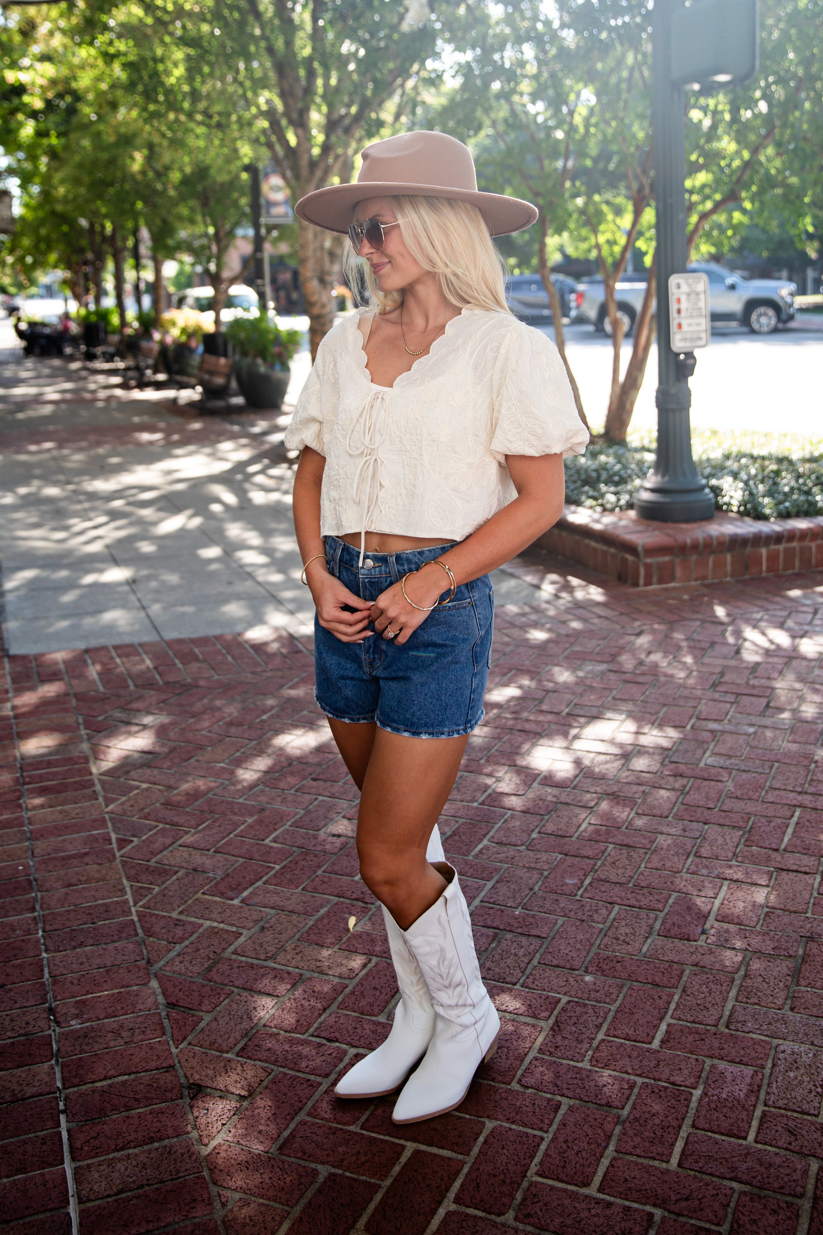 A woman stands on a brick sidewalk outdoors wearing a cream puff-sleeve crop top, denim shorts, white cowboy boots, a beige wide-brim hat, and sunglasses.