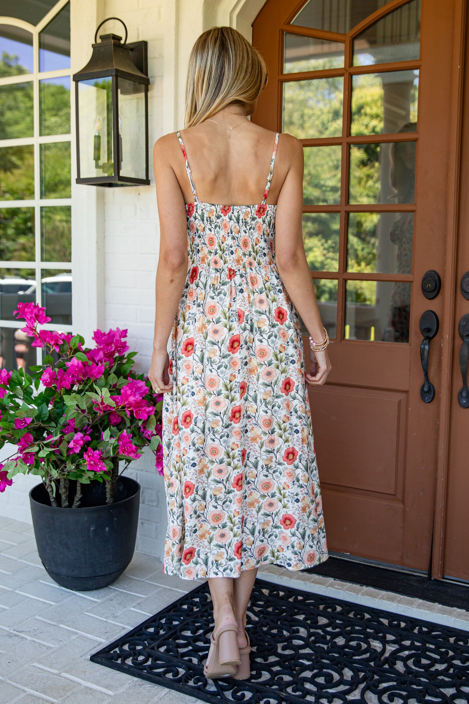 A woman viewed from behind wearing a sleeveless midi dress with a red, orange, and green floral print on a cream background, standing on a porch in front of a brown door.