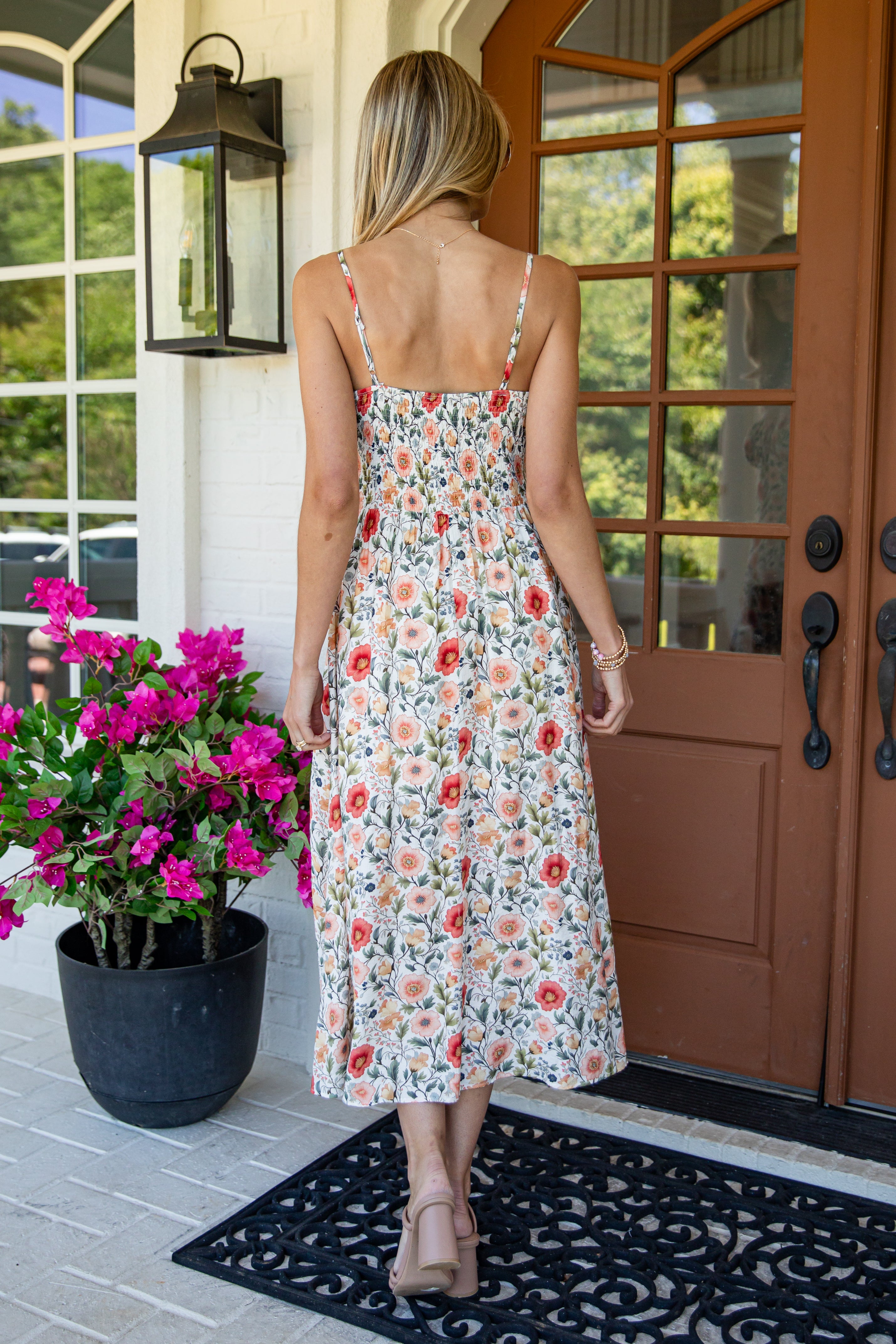 A woman viewed from behind wearing a sleeveless midi dress with a red, orange, and green floral print on a cream background, standing on a porch in front of a brown door.
