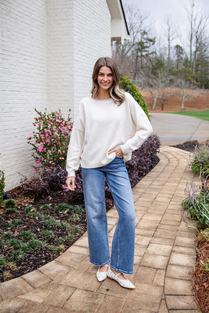 Woman in a white sweater and blue jeans standing on a brick path with plants and flowers in the background.