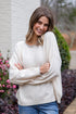 Woman wearing a cream sweater and blue jeans standing outdoors with flowers and a building in the background