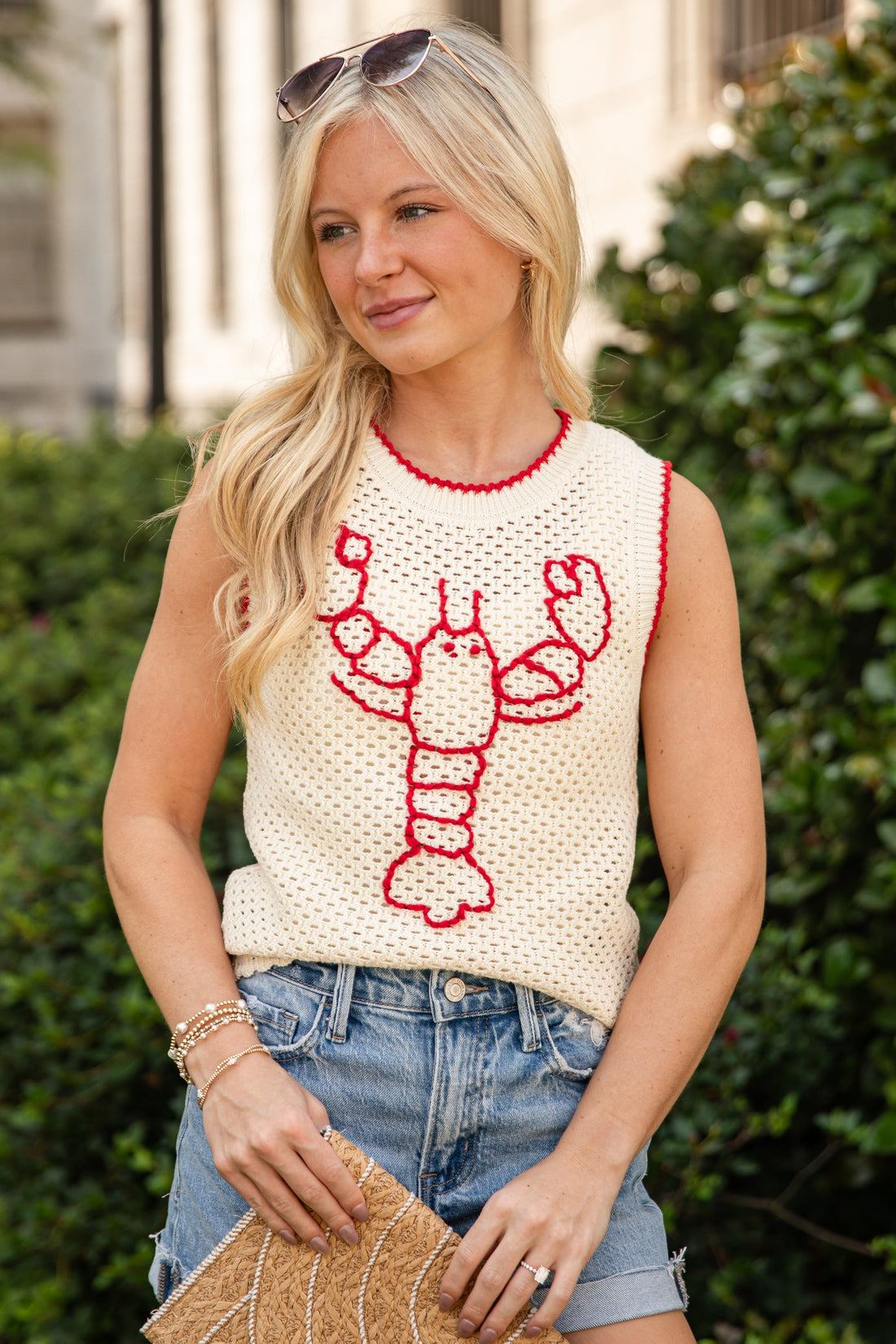 A blonde woman wearing a cream knit sleeveless top with a red lobster graphic and red trim, paired with denim shorts and holding a woven clutch bag, set against a background of green hedges and a building.