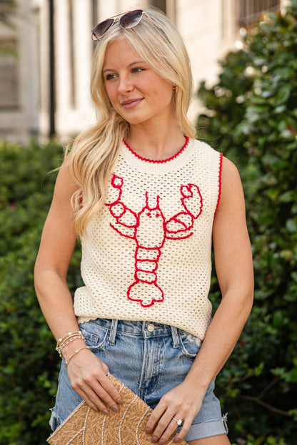A blonde woman wearing a cream knit sleeveless top with a red lobster graphic and red trim, paired with denim shorts and holding a woven clutch bag, set against a background of green hedges and a building.