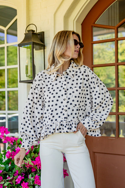 A woman wearing a white long-sleeve blouse with black polka dots and white pants, standing on a porch in front of a brown door and greenery.