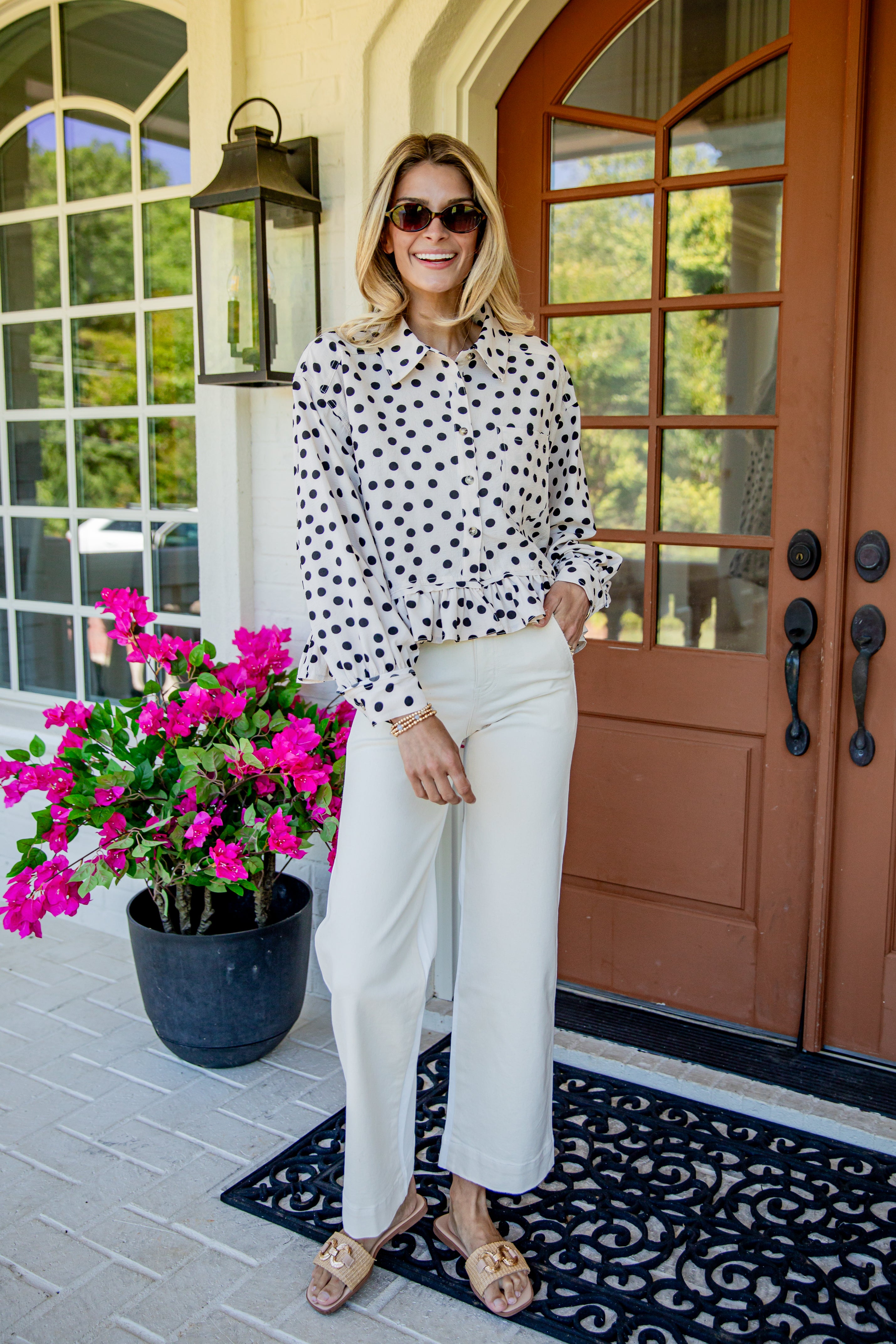 A woman stands on a front porch wearing a white long-sleeve blouse with black polka dots, white wide-leg pants, sunglasses, and tan slide sandals. A potted pink flower sits to her left, and a brown front door is behind her.