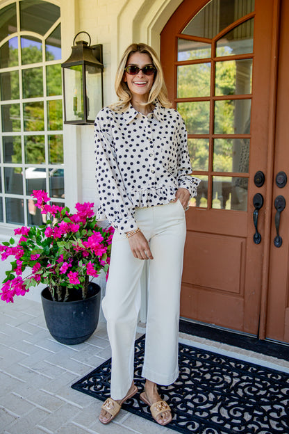 A woman stands on a front porch wearing a white long-sleeve blouse with black polka dots, white wide-leg pants, sunglasses, and tan slide sandals. A potted pink flower sits to her left, and a brown front door is behind her.