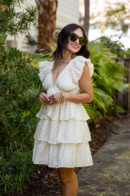 Woman in a white polka dot dress standing outdoors with greenery in the background