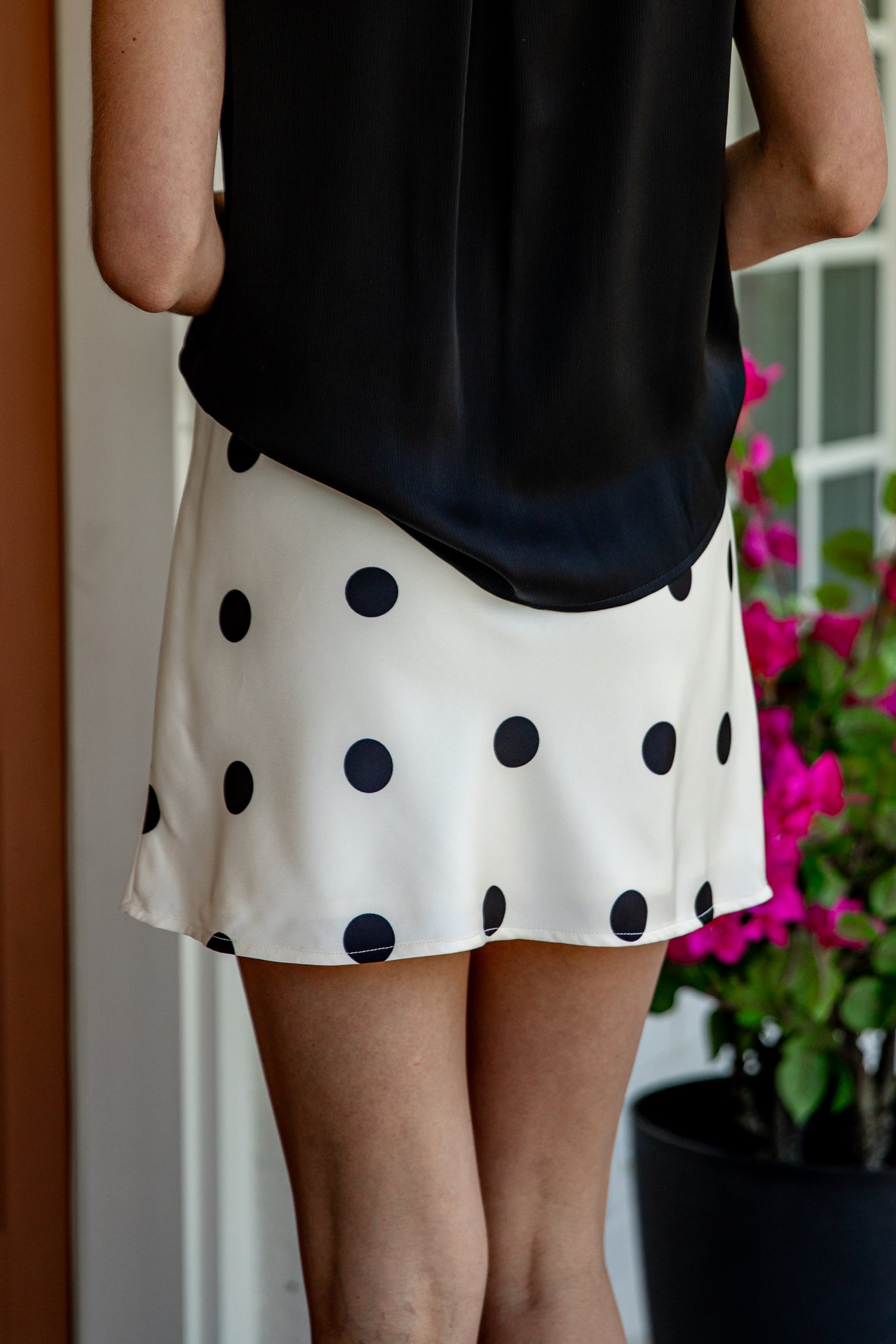 A person wearing a black sleeveless top and a cream A-line skirt with large black polka dots, standing indoors near a potted plant.