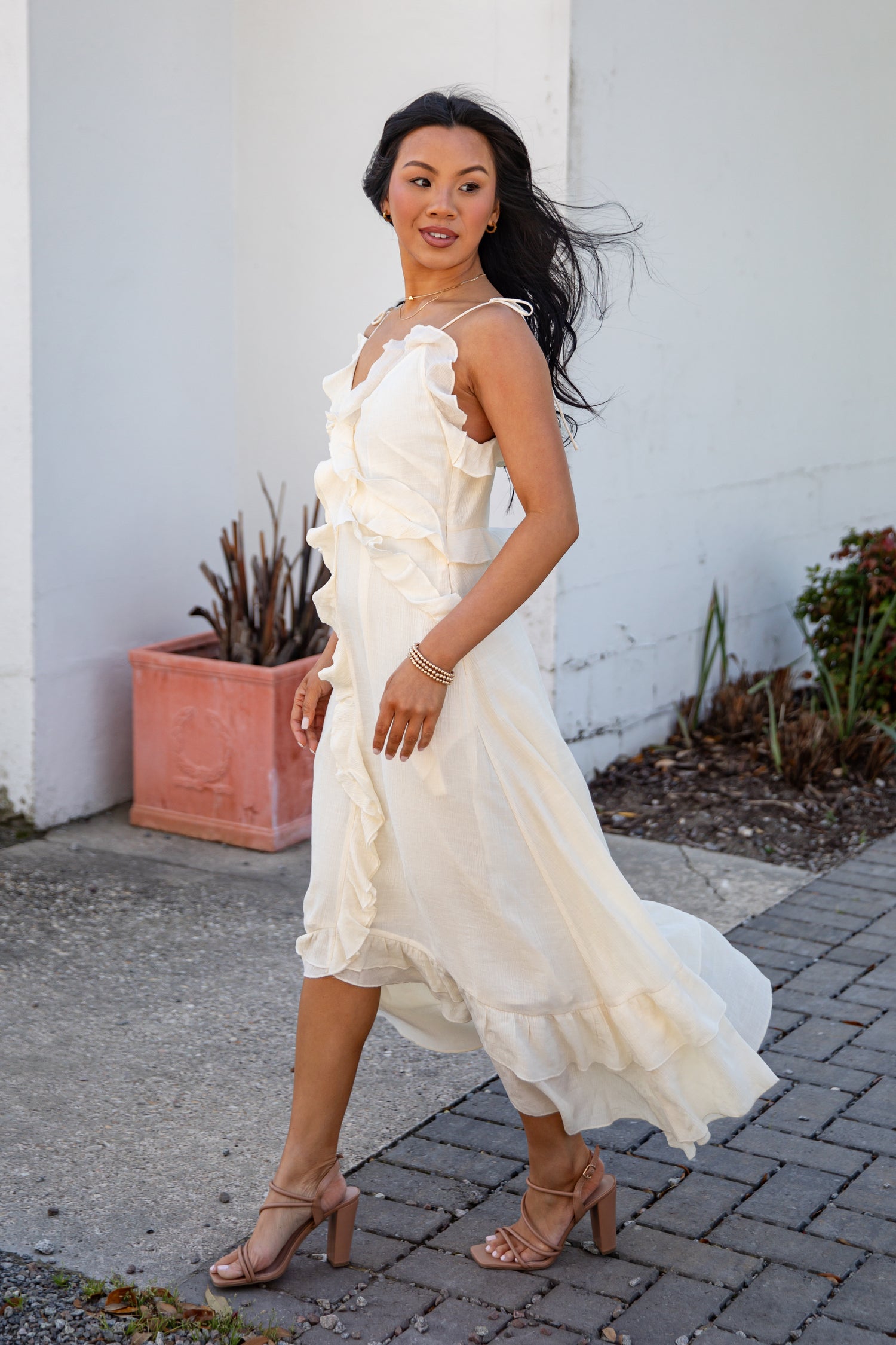 Woman in a white dress standing outdoors against a white wall with plants.