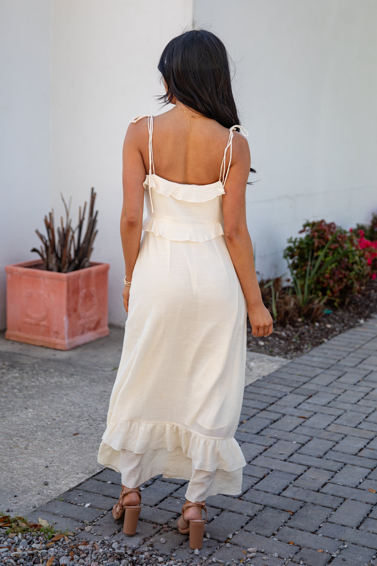 Woman in a white dress walking on a sidewalk with plants and a white wall in the background