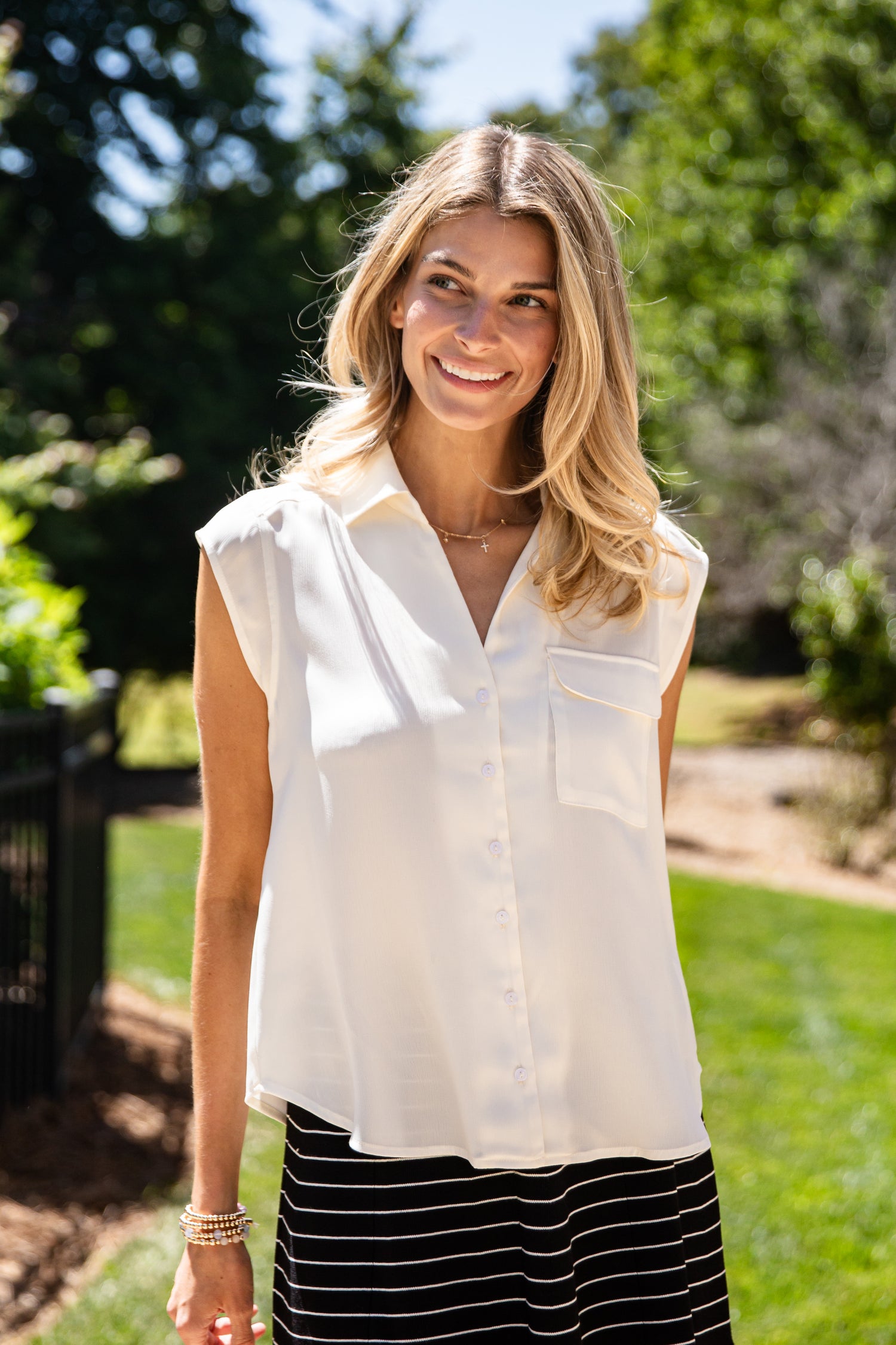 A woman smiling outdoors in a sleeveless white button-up blouse with a collar and chest pocket, paired with a black and white striped skirt.