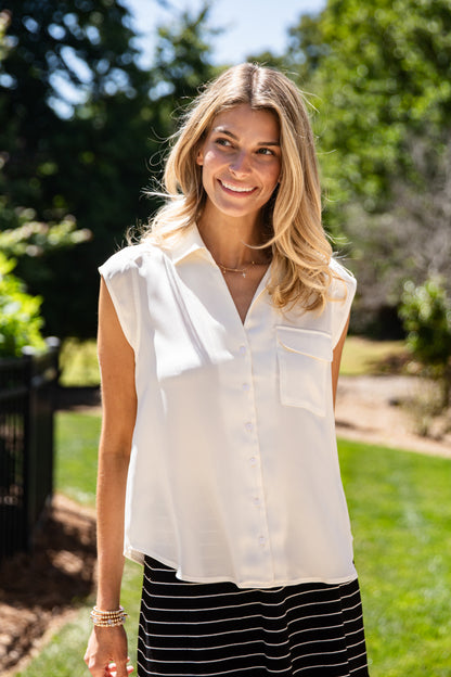 A woman smiling outdoors in a sleeveless white button-up blouse with a collar and chest pocket, paired with a black and white striped skirt.
