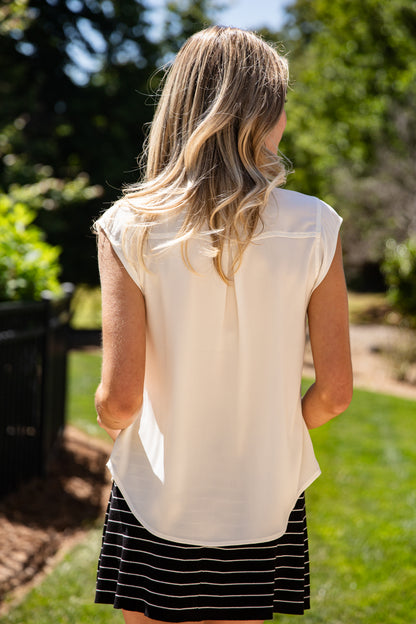 A woman viewed from behind wearing a cream-colored sleeveless blouse and a black and white striped skirt, standing outdoors in a sunny garden setting.