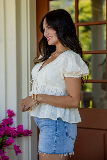 A woman wearing a white short-sleeve lace blouse with puff sleeves and a ruffled hem, paired with a light blue denim mini skirt, standing in front of a porch door.