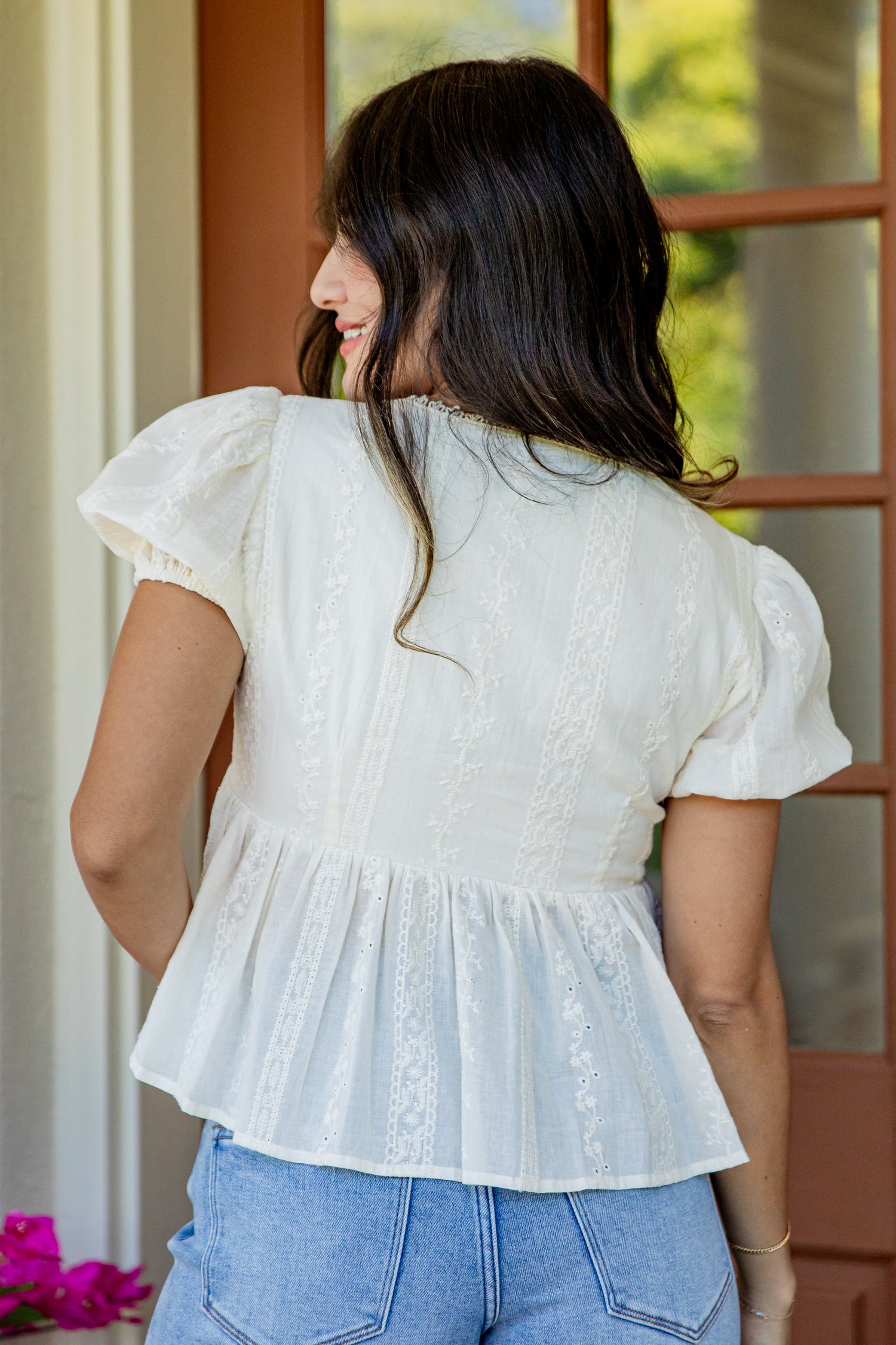 A woman viewed from behind wearing a white short-sleeve peplum blouse with lace detailing, paired with light blue jeans, standing in front of a wooden door.