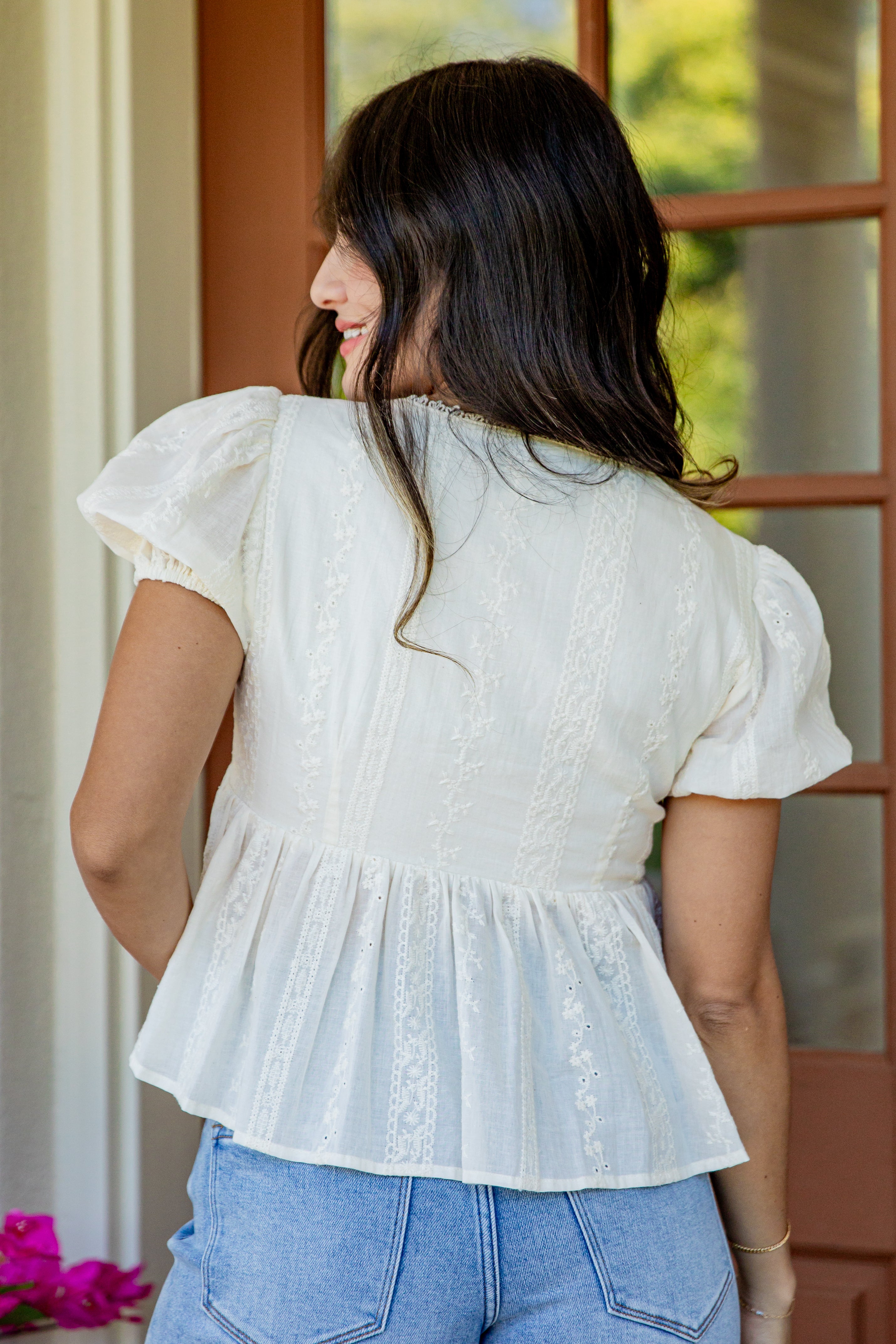 A woman viewed from behind wearing a white short-sleeve peplum blouse with lace detailing, paired with light blue jeans, standing in front of a wooden door.