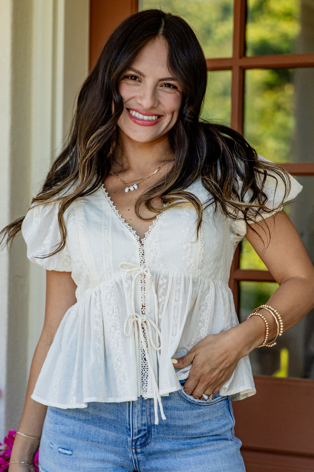 A woman smiling while wearing a white short-sleeve peplum blouse with lace-up front detailing and blue jeans, standing in front of a window.