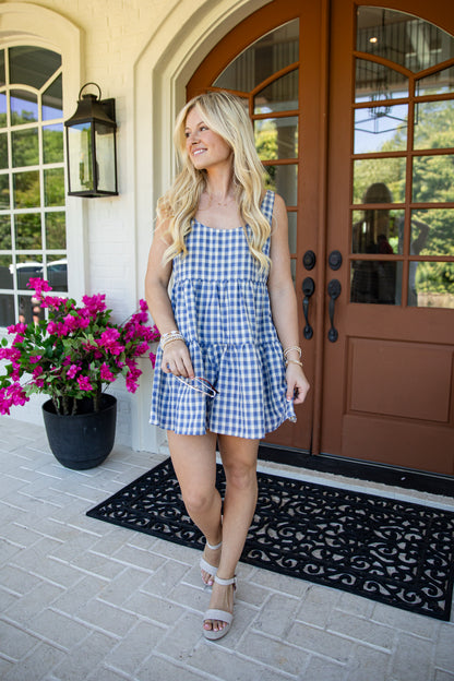 A woman wearing a blue and white gingham tiered romper stands on a front porch in front of brown double doors, with a black doormat and potted pink flowers nearby.