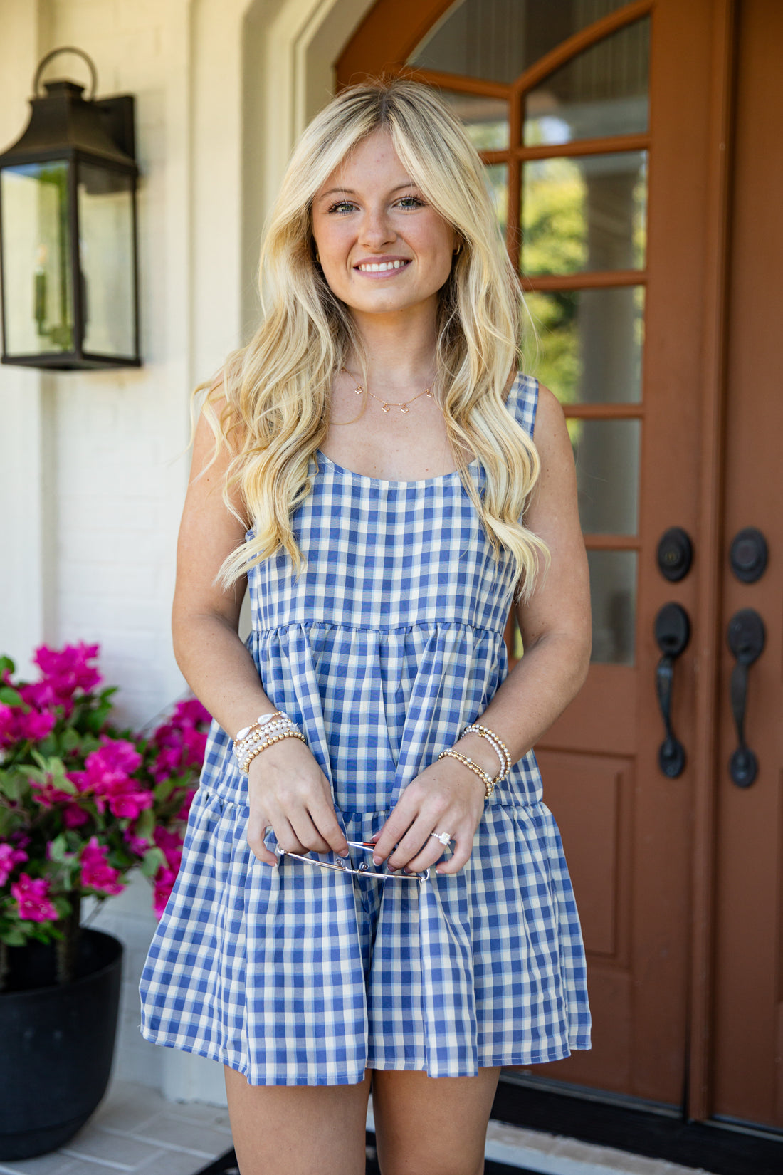 A smiling blonde woman stands on a front porch wearing a blue and white gingham sleeveless romper. She is holding sunglasses, with a black lantern, a potted pink flower, and a brown wooden door in the background.